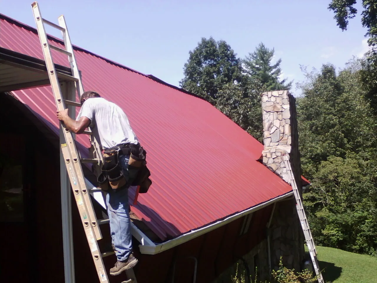 Roofer on a ladder beside a red metal roof and stone chimney — Soffit & Fascia Repair by Roofing Brothers in Branchdale